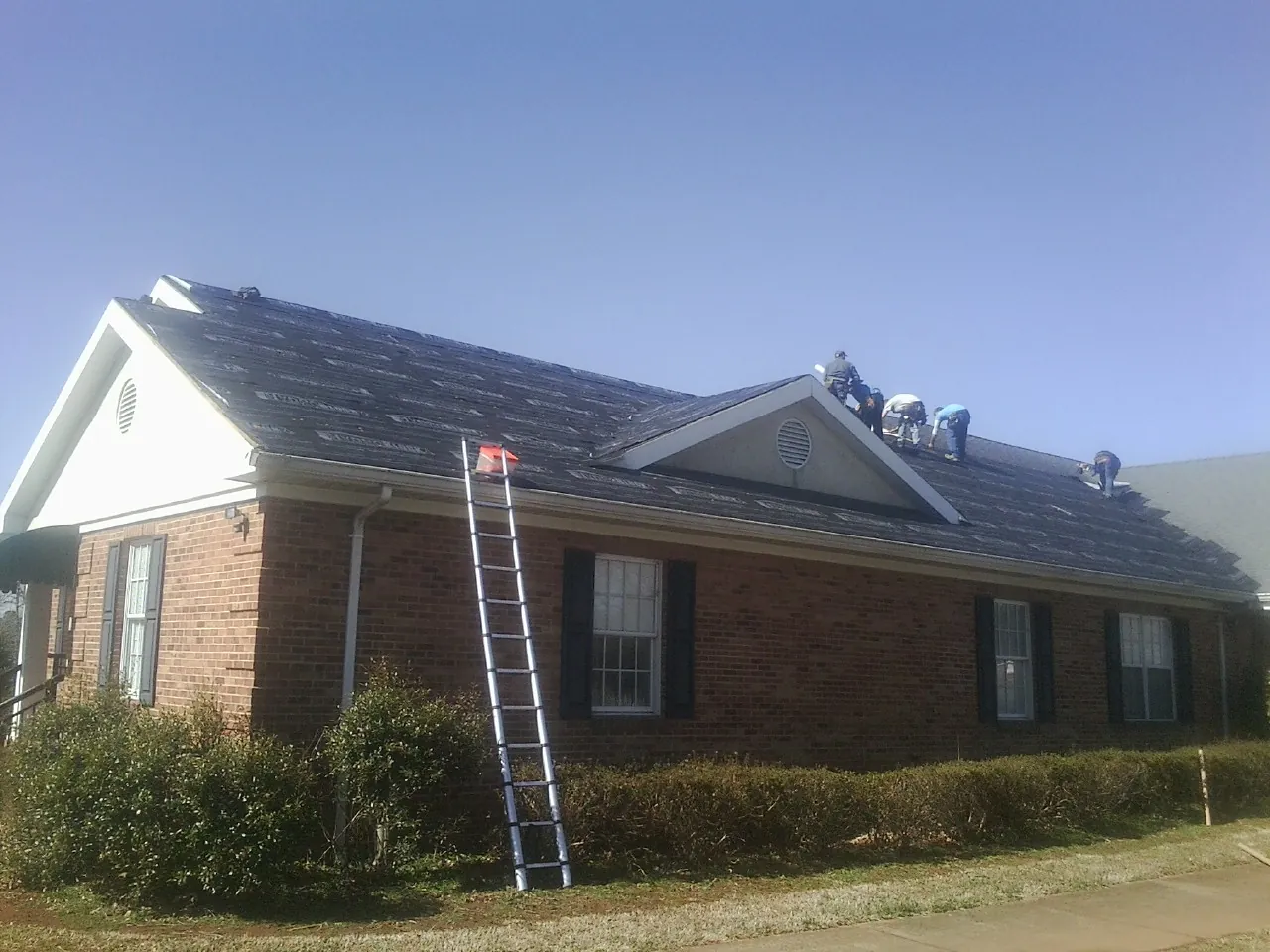 Crew tearing shingles off a brick ranch roof with a ladder set — Emergency Roof Repair work by our crew in Baileyton