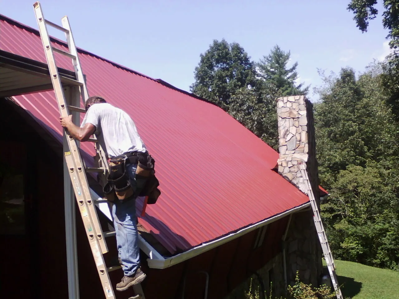 Roofer on a ladder beside a red metal roof and stone chimney — Soffit & Fascia Repair by Roofing Brothers in Baileyton